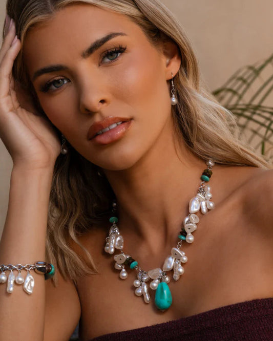 Woman wearing a necklace with turquoise stone and pearl-like beads against a neutral background